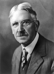 portrait photographique en noir et blanc d’un homme grisonnant vu de face, portant des lunettes, une moustache et une cravate.