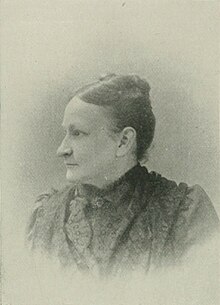 B&W portrait photo of a woman with her hair in an up-do, wearing a dark high-collared blouse.