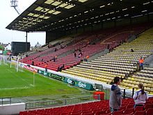 Part of a stadium, consisting of yellow and red seats. A grass football pitch is visible to the left.