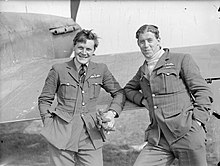 Photographie en noir et blanc de deux hommes souriants en uniforme ; ils se tiennent devant un avion de chasse