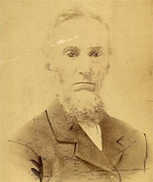 Faded sepiatone portrait photograph of a bearded white man in a suit coat; he is facing and looking into the camera.