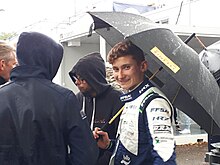 Photographie en couleur d'un adolescent blanc, de trois-quarts gauche, qui regarde l'objectif en souriant, en combinaison de pilote blanche et bleu foncé, tenant un parapluie noir, sous la pluie, accompagné de plusieurs autres hommes.