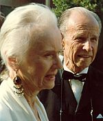 A photograph of an elderly woman seen from the side standing next an elderly man wearing a tuxedo. She is wearing a white dress and an earing.