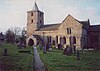 A stone church seen from the south, with a large south transept, and a west tower surmounted by a short recessed lead-covered spire