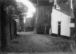Photographie en noir et blanc d'une ruelle encadrée d'un mur de clôture maçonné sur la gauche et par des maisons et un moulin sur la droite.