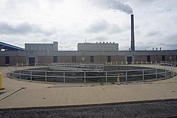 A circular holding tank in the foreground with buildings and a smokestack in the background