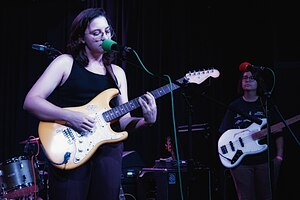 Tucker in a black tank top playing guitar and singing on a stage; Wolfy, their producer, is behind and to the right with a bass.