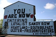 Gable wall of a house covered by a mural with black text in capitals on a blue background. The image of a balaclava-wearing paramilitary holding a machine gun takes up the bottom-left of the wall. Text reads "You are now entering Loyalist Sandy Row / heartland of South Belfast Ulster-Freedom Fighters / Quis separabit". Either side of the text is a red fist.