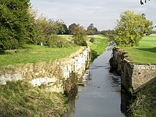 The best preserved of the lock chambers at Alvingham, the retaining walls of brick and stone are intact, but there are no gates. We are looking downstream, away from the weir formed by the upstream sill, which is not visible. On either side of the channel are flat green meadows, and framing those to the edges of the picture are dense woodland. It is a bright sunny day and the trees and grass are a bright green. The data for the picture say it was taken in October 2007, but the vivid green of most of the trees, and particularly one just on the right of the lock, looks like spring.