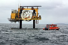 Photo taken at sea of the OpenHydro turbine, raised above the water supported between 2 metal piles. There is a walkway around the turbine for maintenance access, and a small orange boat nearby. The turbine has an outer and inner ring, with 10 blades between.