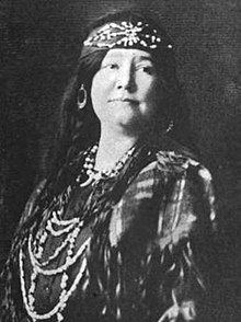 A woman with fair skin and dark hair; she is wearing a beaded headband, braids, beads, a fringed shawl.