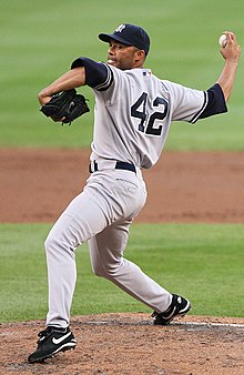 A right-handed Hispanic baseball pitcher, wearing a grey uniform with the lettering "NEW YORK" across it, with his body facing the right as he prepares to throw a baseball.