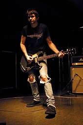 A male musician, Steve Albini, with an electric guitar strapped over his shoulders in a playing position. He is wearing blue jeans with large rips in both knees.