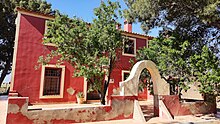 Entrada de la casa con un arco, rodeado a la izquierda por un almendro y a la derecha por un pino. Al fondo la casa, de color rojo con dos plantas. Se aprecia que tiene un gran número de ventanales.