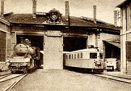 Photographie en sépia d'une locomotive à vapeur et d'un autorail devant une rotonde ferroviaire.