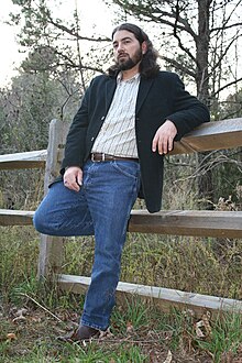 Ronnie Penque, wearing blue jeans and a jacket and leaning against a wooden fence