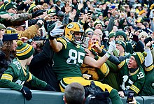 Packers tight end Robert Tonyan jumping on top of the wall around Lambeau Field, leaning in to the fans situation in the stands, after scoring a touchdown