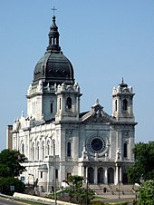 A large white basilica with a black roof stands on a sunny day.