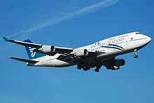  An Air New Zealand 747-400 with its landing gear down and flaps down.  The aircraft is mostly white and blue aircraft and in-flight against a blue sky. On each of the two wings are two engines.