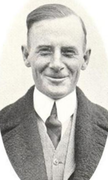 A smiling white man wearing a suit and tie; he is cleanshaven and he has short hair with a receding hairline
