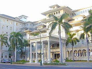 Starwood Sheraton Moana Surfrider Hotel, with a front side made of white stone decorated with arches and round pillars, with palm trees in front of the building.