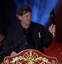 middle-aged white male wearing a dark suit, standing at a podium, holding trophy, looking left of camera