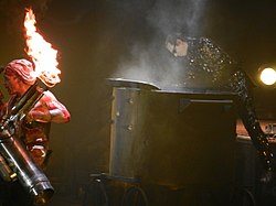 Till Lindemann iført et slakterkostyme holder en flammekaster, imens Flake Lorenz går ut av en stor potte iført glitterklær.
