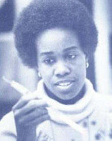 A young Black woman with an Afro hair style, wearing a turtlenece sweater and holding a piece of laboratory equipment