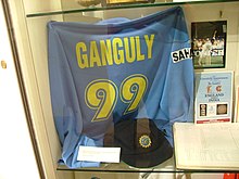 A blue colored t-shirt displayed at a store window. The t-shirt has the words "Ganguly" and the number 99 below it, both in yellow color. Beside the t-shirt, a picture and an open book is visible.