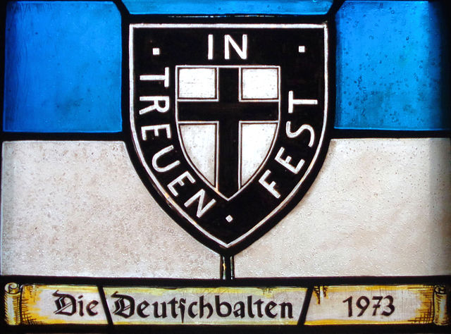 Farbfotografie eines Glasfensters mit blauem und weißem Querstreifen, auf dem ein schwarz-weißes Wappen mit einem schwarzen Kreuz und der Aufschrift „In Treuen fest“ steht. Auf einer ausgerollten Pergamentrolle im unteren Fensterbereich steht „Die Deutschbalten 1973“.