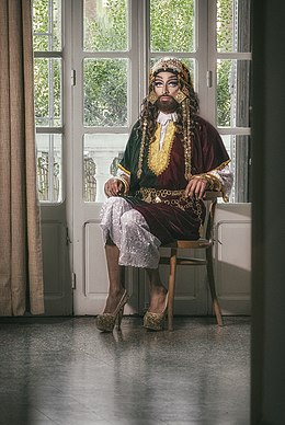 Een dragqueen met een baard, traditionele kleding en schoenen met hakken zit in een stoel