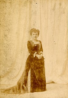 A sepia-toned full-length photograph from 1890, of a woman in a gown.
