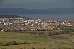 Multiple buildings including terraces, detached houses and blocks of flats. In the foreground are fields and in the background water and then hills.