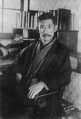 Photo noir et blanc d'un homme vu de trois quarts, assis sur une chaise, devant un bureau et une étagère supportant des livres (en haut, à droite)