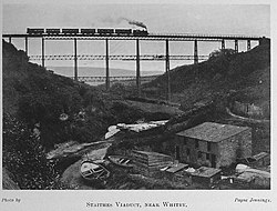 A black and white image of a tall, narrow, metal viaduct, crossing a steep, narrow s-shaped valley, with a small beck and fishing boats on the valley floor