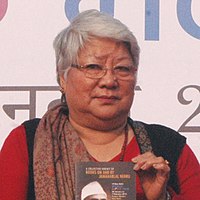 Photo en couleurs d'une dame indienne à cheveux blancs et lunettes, présentant un livre.