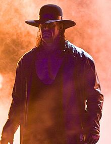 A man with long dark hair, wearing a black wide-brimmed hat, black leather jacket, and black wrestling tights.