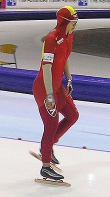 Une patineuse en combinaison rouge, portant également un brassard blanc et des lunettes. Elle se tient sur le pied droit et est complètement debout.