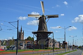 Photographie en couleurs d'une route, un moulin à vent, un clocher d'église et des bâtiments évoluant sur sa gauche.