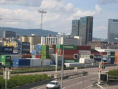 Foto einer Stadt mit einer mehrspurigen Straße im Vordergrund, einem Lagerplatz mit Schiffscontainern dahinter und Hochhäusern im Hintergrund