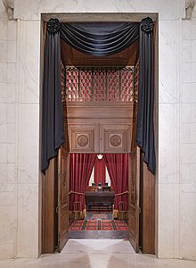 Двери зала заседаний Верховного суда задрапированы черным. Сквозь открытую дверь видно сиденье Гинзбурга и скамейку перед сиденьем, каждое из которых также задрапировано черным.