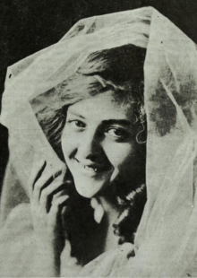 A smiling young white woman with her head loosely covered by white gauzy fabric
