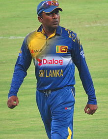 Portrait of dark skinned man, wearing blue and yellow Sri Lanka cricket team kit with cap. Cricket field in the background.
