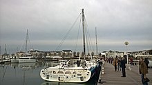 Vu de l'arrière, dans un port, sans les voiles, bateau au mât remplacé par la bôme cassée en deux et réassemblée durant la course.