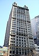 Ground-level view of a 30-story building with a tan exterior and a square cross-section; spires sit atop the roof of the building at each corner