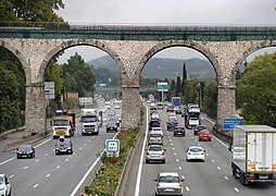 Photographie en couleurs représentant une autoroute à deux fois trois voies passant sous deux arches du pont et parcourue par des voitures et des camions.