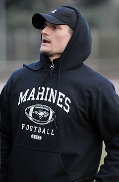 Candid waist-up photograph of Olivo wearing a black hooded sweatshirt bearing the text "Marines football" and a black hat with an Under Armour logo