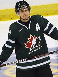 Half-length portrait of hockey player in black Canada uniform on the ice. His lips are pressed together ruefully and he looks into space as if thinking.