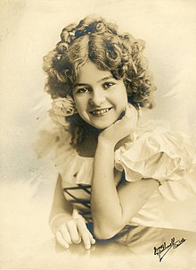 A smiling white girl with curled sandy hair, posed in a white ruffled dress