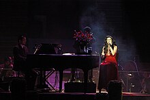 A woman in a red dress sings into a hand-held microphone while sitting on a chair beside a piano during a concert